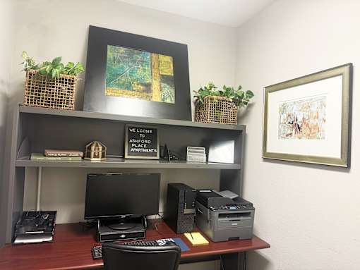 A black monitor and a black printer on a desk with a sign that says "We Welcome Ashford Place Apartments".