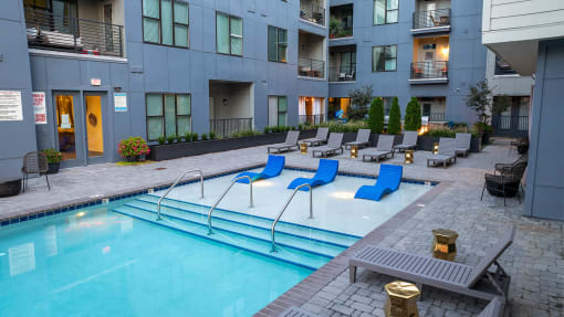 an apartment pool with blue lounge chairs in front of an apartment building