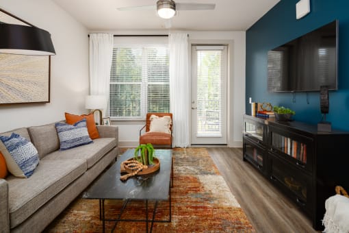 2-Bedroom Apartments in Belmont, NC- The Banks Apartments - Living Room with Wood-Inspired Plank Flooring, Area Rug, Sofa, Coffee Table, Armchair, Ceiling Fan, Storage Cabinet, Wall-Mounted TV, Window, and Glass Door with Access to Private Balcony.