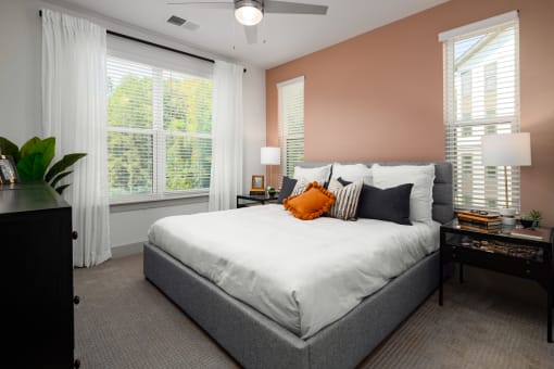 One-Bedroom Apartments in Belmont, NC - Banks at Mt. Holly - Bedroom with Carpeted Floor, Large Bed, Nighstands with Lamps, Dresser, Large Windows with Blinds and Curtains, and Ceiling Fan