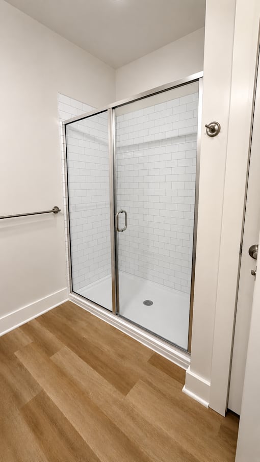 A white tiled shower with a glass door is in a room with wood flooring.