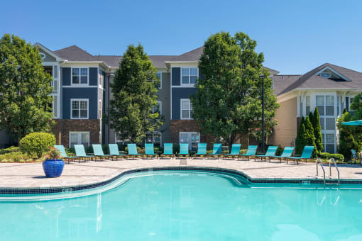 Chapel Hill, NC, Luxury Apartments with In-Unit Washers and Dryers - The Reserve at Meadowmont - Resort-Style Swimming Pool with Sundeck, Lounge Chairs, Surrounding Trees, and Apartment Buildings in the Background.