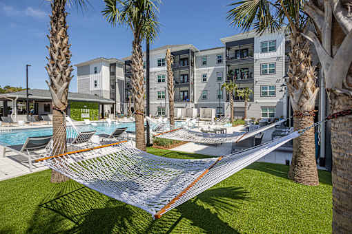 Panama City, FL, Apartments Near Santa Rosa Beach - The Tyde Apartments - Hammock Lounge With Green Turf, Palm Trees, Lounge Chairs, and Apartment Buildings Under Blue Sky.
