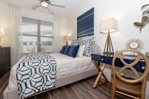 Pet-Friendly Luxury Apartments in Atlanta, GA - 1105 Town Brookhaven - Bedroom with Hardwood Floor, Bed, Desk with Chair and Lamp, Ceiling Fan, and Large Window.