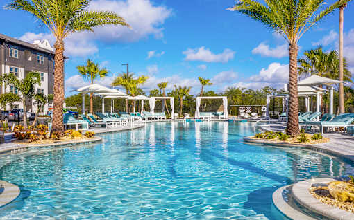 Luxury Apartments in Sarasota, FL With In-Unit Washer And Dryer - Botanic Waterside - Resort-Style Swimming Pool With Spacious Sundeck, Lounge Chairs, Umbrellas, Cabanas, And Palm Trees.