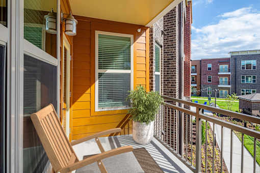 2-Bedroom Apartments in Mt. Juliet, TN - The Enclave at Providence - A Balcony with an Orange Siding, Large Windows, Light Fixture, Wooden Lounge Chair, Brown Rug, Metal Railing, and Potted Plant.