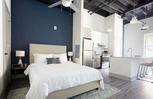 A bedroom with a large bed and a kitchen area in the background.