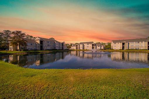 A serene landscape with a lake, grass, and buildings in the background.