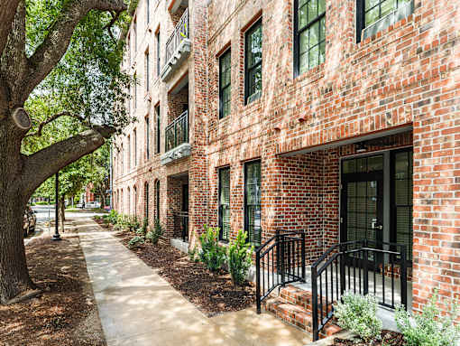 A tree-lined brick walkway leads to a building with a black railing.