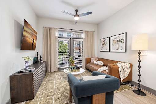 Duluth, GA, Apartments near Mercedes-Benz Stadium - District at Duluth - Living Room with Hardwood-Style Flooring, a Ceiling Fan, a Mounted TV, a Couch, and Doors to a Balcony.