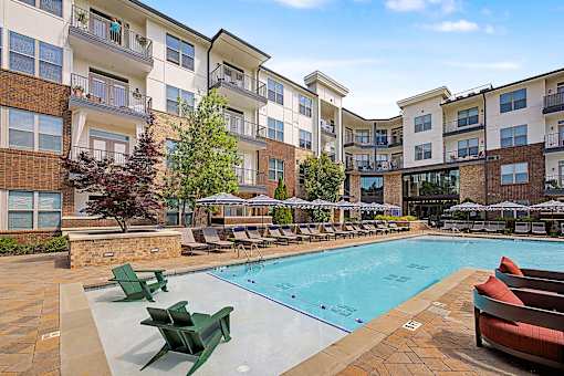 A large swimming pool surrounded by lounge chairs and umbrellas in front of apartment buildings.