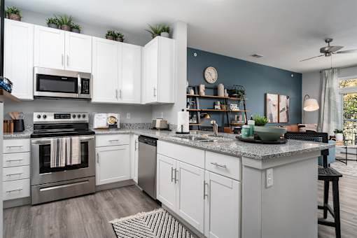 Apartments for Rent in Alexandria, KY - Elevation Apartment Homes - Kitchen with 9-Foot Ceiling, Wood-Style Flooring, White Shaker-Style Cabinetry, Stainless-Steel Frigidaire Appliances, Granite Countertops, Kitchen Tools, Decorative Plants, Eat-in Kitchen with Breakfast Bar, Bar Stool, and a View of the Living Area and Balcony Doorway.