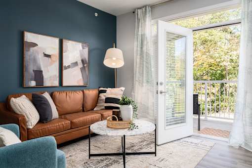 Apartments in Alexandria, KY, with In-Unit Washer and Dryer - Elevation Apartment Homes - Living Room with 9-Foot Ceiling, Wood-Style Flooring, Large Rug, Brown Couch, Blue Armchair, Round Center Table, Floor Lamp, 2 Large Wall Arts, Home Decor, Floor Length Curtains, and an Opened Doorway to the Balcony.