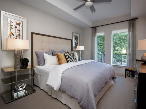 Dog-Friendly Luxury Apartments In Atlanta, GA - Venue Brookwood - Spacious Bedroom With Carpet Flooring, Large Bed, Multi-Level Nightstands With Lamps, Ceiling Fan, Dresser, Chair, And Large Windows.