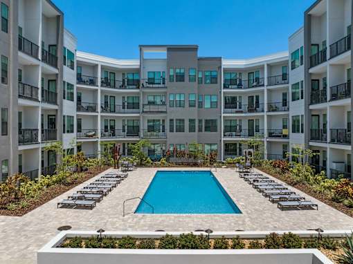 A large swimming pool surrounded by lounge chairs in front of apartment buildings.