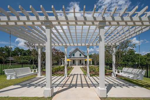 A white pergola structure is in the foreground of a park with benches and a building in the background.