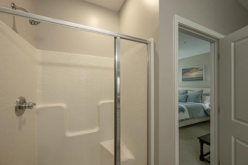 A white bathroom with a glass shower door and a bedroom visible through the door.