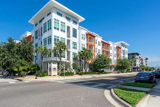 A modern multi-story building with a white and orange facade is situated on a street corner with cars parked along the curb.