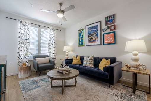 One-Bedroom Apartments in Tampa, FL - Havana Square - Living Room with Vinyl Plank Flooring, Area Rug, Sofa, Armchair, Coffee Table, Side Tables with Lamps, Wall Art, Ceiling Fan, and Large Window with Coverings.