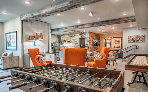 A foosball table is in the middle of a room with orange chairs and a stone wall.