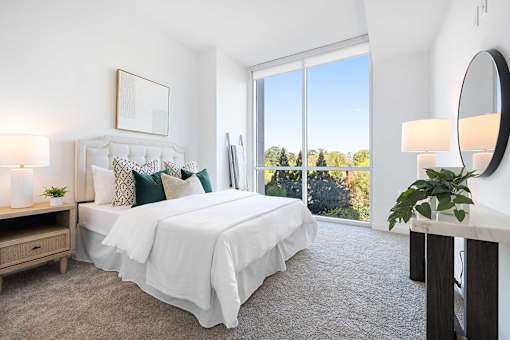 A bedroom with a large bed, a nightstand, a lamp, a mirror, and a window with a view of trees.