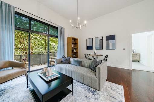 A living room with a grey couch, a coffee table, and a large window with a view of trees.