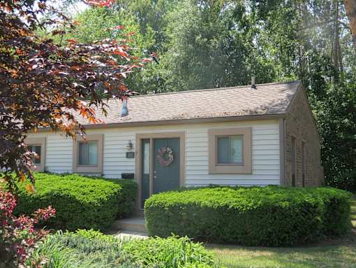 A small white house with a green door and window is surrounded by greenery.