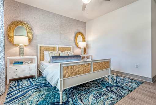 1-Bedroom Luxury Apartments In Sarasota, FL - Botanic Waterside - Bedroom With Wood-Finish Flooring, Colorful Area Rug, Large Bed, Nightstands With Lamps, Wall-Mounted Mirror, Accent Wall, And Ceiling Fan.