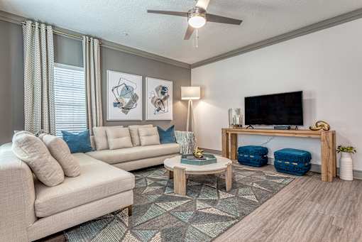 2-Bedroom Luxury Apartments In Sarasota, FL - Botanic Waterside - Living Room With Wood-Finish Flooring, Area Rug, Sectional Couch, Coffee Table, Stools, Display Shelf, Wall-Mounted TV, Crown Molding, And Window.