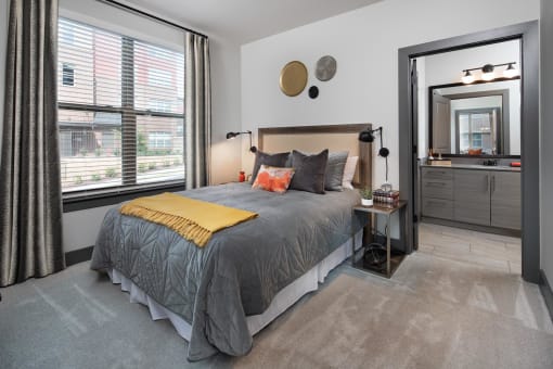 Duluth, GA, Apartments Near Stone Mountain Park - District at Duluth - Bedroom with Carpeted Floor, Bed, Nightstands with Lamps, Oversized Window, and Attached Bathroom.