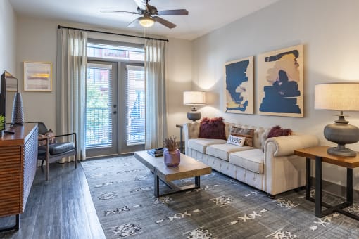 Duluth, GA, Apartments Near Georgia Aquarium - District at Duluth - Living Room with Hardwood Floor, Area Rug, Sofa, Coffee Table, Side Tables with Lamps, Wall Art, Storage Cabinet, TV, Armchair, Ceiling Fan, and Glass Doors to Private Balcony.
