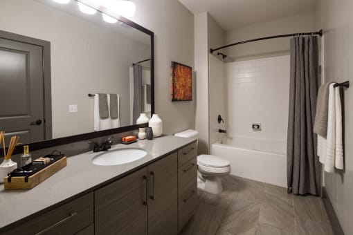 Duluth, GA, Apartments Near Mercedes-Benz Stadium - District at Duluth - Bathroom with Tile Floor, Garden-Style Tub, Quartz Vanity Countertop, Large Mirror, and Wood-Tone Cabinets.