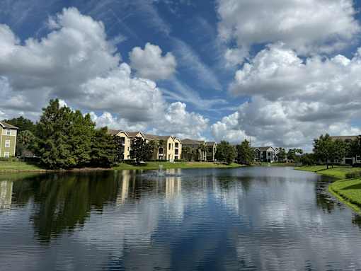 A serene lake surrounded by houses and trees under a blue sky with clouds.
