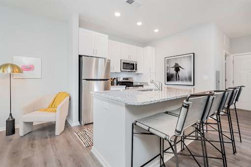 Luxury Apartments in Louisville, KY, with In-Unit Washer and Dryer - The Laurel Apartments - Kitchen With White Cabinetry, Stainless Steel Appliances, Granite Island Seating, And A Cozy Accent Chair.