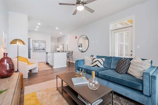 Lebanon, OH, Luxury Apartments For Rent - Founders Landing - A Modern Living Area With A Blue Velvet Sofa, A Gold Floor Lamp, And A White Kitchen Island In An Open-Concept Layout.