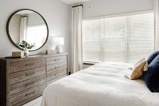 A bedroom with a white bed and a wooden dresser with a round mirror above it.