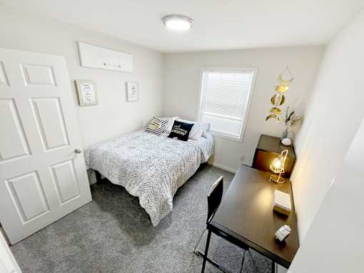 Harrisonburg, VA, Fully Furnished Student Apartments - The Mill - A Bright, Minimalist Bedroom With A Rustic Wood Dresser, A Large Window, And A Bed With Neutral-Toned Linens.