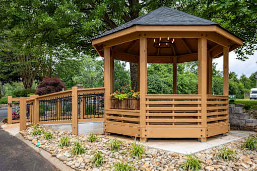 A wooden gazebo with a black roof is surrounded by a metal fence and a gravel area with plants.