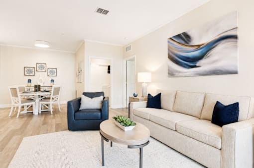 A living room with a white couch, a blue chair, a dining table with chairs, a lamp, and a painting on the wall.