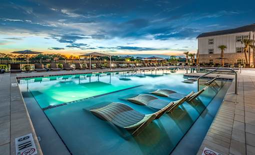 Luxury Apartments In Jacksonville, FL, With In-Unit Washer And Dryer - The Clarke At Kendall Town - A Large Swimming Pool At Sunset Featuring In-Water Lounge Chairs.
