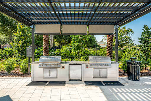 Two grills are under a roof in a sunny outdoor area.