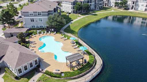 A large swimming pool with a small waterfall and a hot tub is surrounded by a grassy area and a paved walkway.