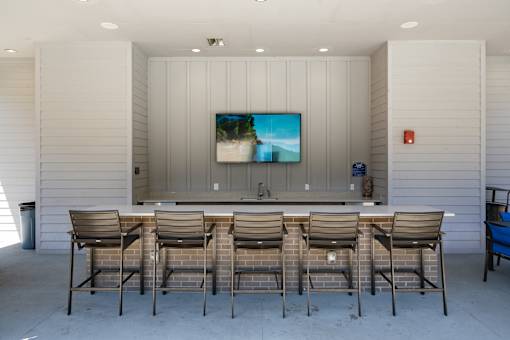 A television is mounted on a wall above a counter with four chairs in front of it.