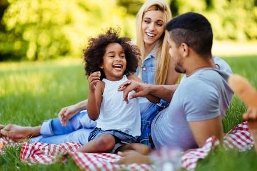A family of three is sitting on a blanket in a park.