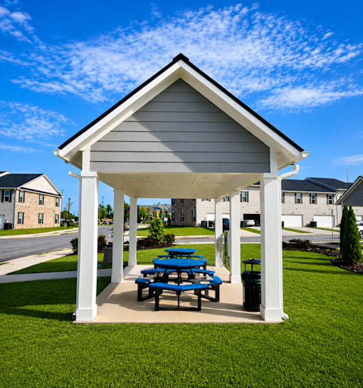A pavilion with a white roof and grey walls is surrounded by a grassy area and a blue picnic table.