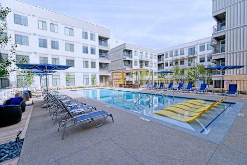 Studio Apartments in Downtown Memphis, TN - The Rise - Outdoor Resort-Style Pool with Surrounding Lounge Seating, Blue Umbrellas, Separate Lounge Areas with Wicker Tables and Seating, and an Exterior View of the Property Building.