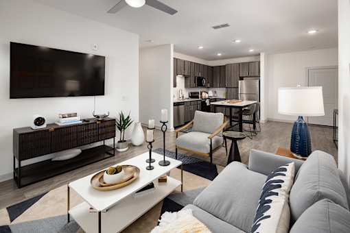 Luxury Apartments in Ridgeville, SC, with In-Unit Washer and Dryer - Preserve at Ridgeville - Living Room with Wood-Inspired Flooring, Area Rug, Couch, Armchair, Coffee Table, Side Table with Lamp, Ceiling Fan, Media Console, Wall-Mounted TV, and View of Kitchen Area.