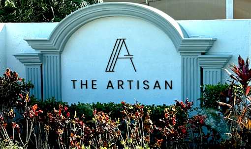 A white sign with a black letter A and the words "The Artisan" in front of some plants.