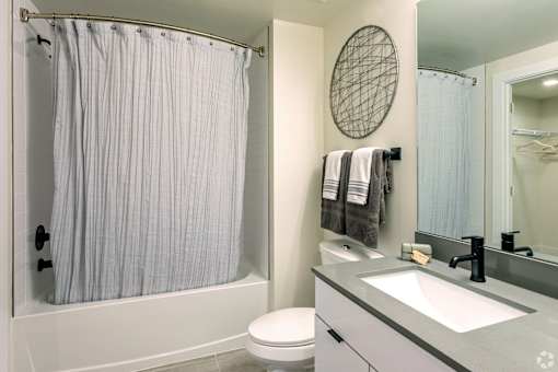 A bathroom with a white shower curtain, a round mirror, and a white toilet at Saba Pompano Beach, Pompano Beach, 33062