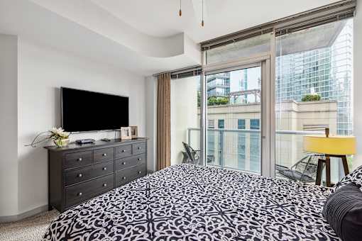 A bedroom with a patterned bedspread, a television, a dresser, and a window with a view of a cityscape.